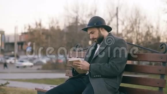 有创意的年轻作家戴着帽子穿着外套坐在城市街道上用钢笔为他未来的书写笔记视频的预览图