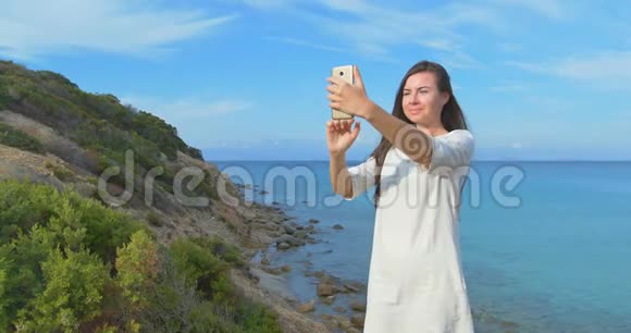 年轻漂亮的女人用她的智能手机在海上拍自拍照片日落时女性站在海边清楚视频的预览图