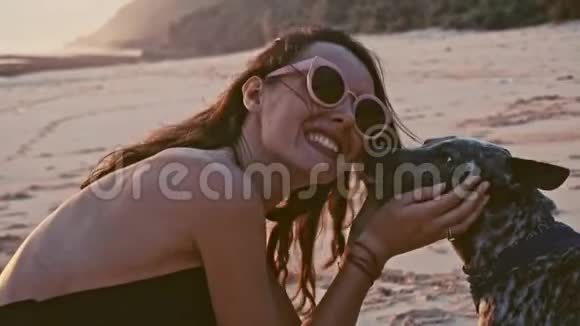 年轻的无忧无虑的女人戴着太阳镜在海滩上玩狗在惊人的日落海洋暑假概念视频的预览图