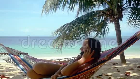 迷人的黑发女人在棕榈树旁边的热带海滩上在手机上聊天在吊床上放松美丽视频的预览图