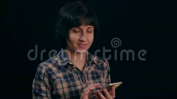一位穿着格子衬衫的漂亮年轻女士在黑色背景下使用智能手机视频的预览图