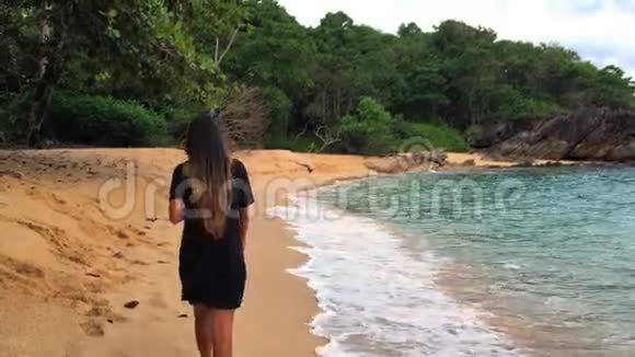 穿着黑色衣服的年轻女子在秘密海滩沿着海边散步喝着咖啡带走了慢速高清视频的预览图