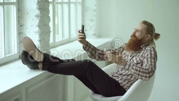 有胡子的年轻时髦商人在智能手机上聊天坐在椅子上双腿搭在椅子上视频的预览图