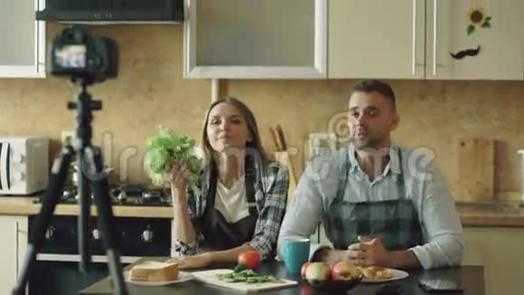 年轻漂亮的情侣在厨房里用dslr摄像机拍摄有关烹饪的视频食物博客视频的预览图