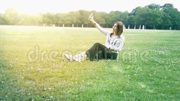 美丽的女孩坐在草坪上在智能手机上自拍视频的预览图