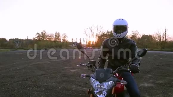一个穿着黑色皮夹克和白色头盔的年轻人坐在摩托车上在秋日日落前开灯视频的预览图