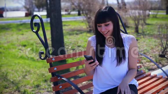 一个穿着白色衬衫的东方美女坐在公园里的长凳上手里拿着一部智能手机非常漂亮视频的预览图