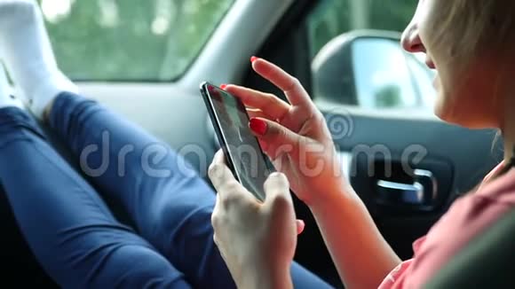 乘客女孩正坐在汽车的前座上在电话上留言把脚踩在车上视频的预览图