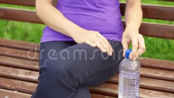 微笑的年轻女人享受大自然坐在绿色公园的长椅上喝着瓶子里的水视频的预览图