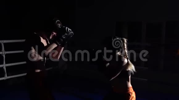 一个戴着头盔和拳击手套的女孩在黑暗中在拳击场上打的家伙慢动作视频的预览图