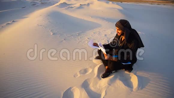 英俊的阿拉伯男性建筑师看着文件在温暖的夏日傍晚坐在沙漠中的沙滩上视频的预览图
