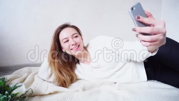 美丽的女孩在智能手机上拍了一张令人难忘的自拍的相机在新年的心情中躺在地板上明亮的房间里的格子布上视频的预览图
