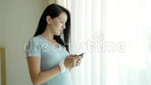 使用智能手机的年轻女性肖像高加索模特站在窗口用她的智能手机视频的预览图