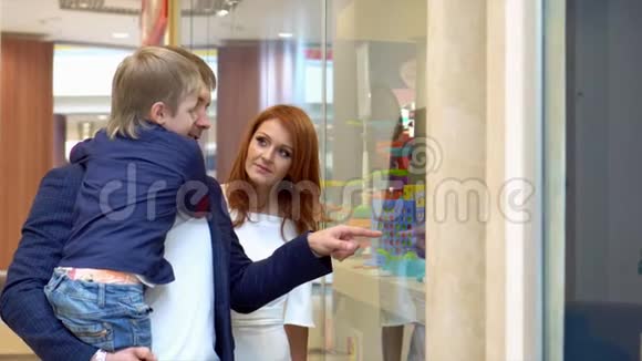 带着父母的小男孩看着一个带玩具的橱窗这个男孩为圣诞节挑选礼物一头金发视频的预览图