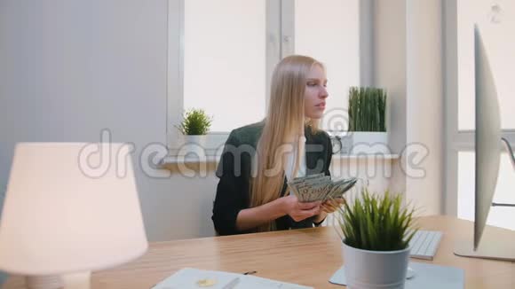 在办公室庆祝成功的女人优雅的金发女性坐在工作场所手里拿着一捆现金看上去很漂亮视频的预览图