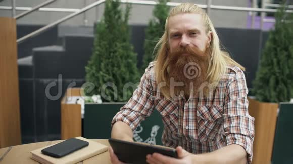 留胡子的年轻人带着平板电脑坐在户外的咖啡馆里微笑放松和休息视频的预览图