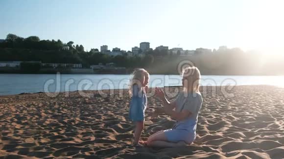 美丽快乐的金发妈妈和女儿在日落时在海滩上拥抱和说话视频的预览图