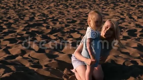 美丽的金发妈妈和女儿在日落时在海滩上拥抱和说话视频的预览图