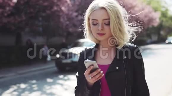 漂亮的金发女孩穿着休闲装走在樱花巷梦幻般的微笑使用她的手机好好享受视频的预览图