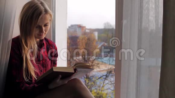 美丽的金发女郎正在看书坐在窗台上视频的预览图