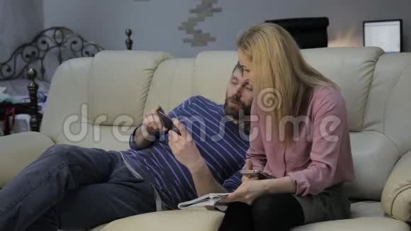 金发学生读她的笔记她的男朋友坐在沙发上用智能手机视频的预览图