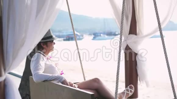 快乐的女人戴着帽子戴着太阳镜在一个热带海滩上的木秋千上度过暑假慢慢慢慢视频的预览图