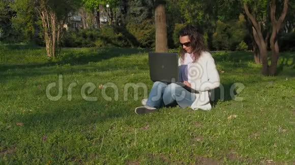 在大自然中工作的女人公园里的女孩拿着电脑坐在草地上戴墨镜的年轻女子视频的预览图