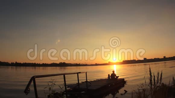 剪影一个小孩和一个女人在日落时坐在水边的码头上视频的预览图