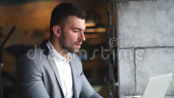 一个穿着灰色西装和白色衬衫的年轻商人在咖啡馆的笔记本电脑上工作视频的预览图