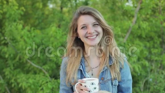 可爱的长发女孩笑着手里拿着一个杯子在绿色灌木丛的背景下穿牛仔裤的漂亮金发女孩视频的预览图