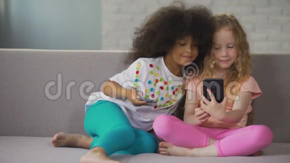 多种族姐妹坐在沙发上在家里的智能手机上自拍视频的预览图