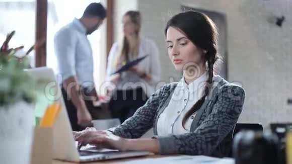 漂亮的黑发女人在轻型办公室的笔记本电脑里打字其他工人可以在背景中看到视频的预览图