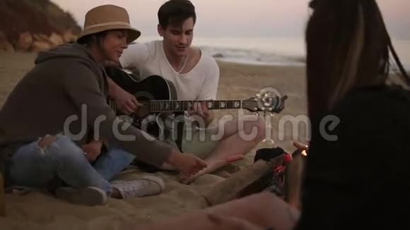 年轻人正和朋友一起坐在海滩上的火炉旁弹吉他他女朋友烤香肠视频的预览图