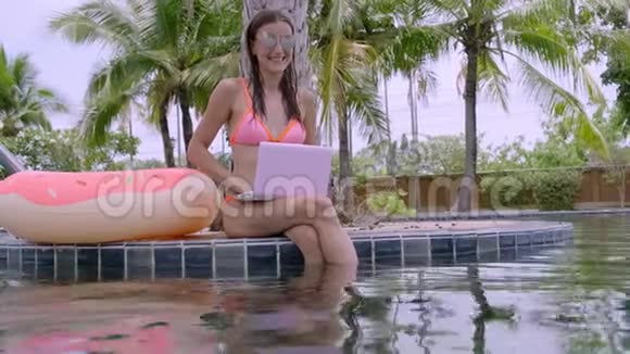 一个年轻的女人坐在泳池边手里拿着一台微型电脑选择性聚焦视频的预览图