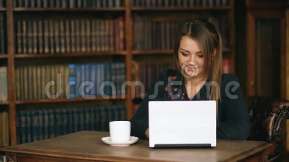 带着笔记本电脑的年轻微笑女子在图书馆通过室内网络摄像头与朋友交谈视频的预览图