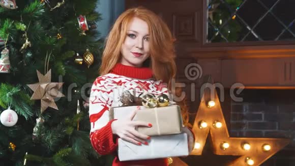 可爱的红头发女孩穿着红色毛衣靠近圣诞树手里拿着礼物视频的预览图