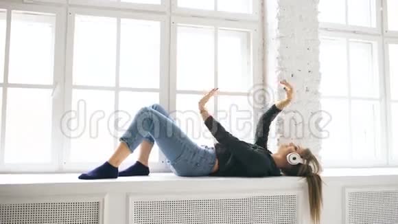 带着耳机听音乐跳舞的年轻女子躺在室内的窗户上视频的预览图