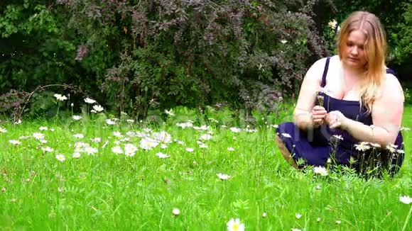 一个穿着蓝色萨拉芬衣服的女孩收集坐在绿草上的野花视频的预览图