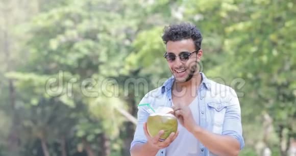 西班牙男人在棕榈树上喝椰子鸡尾酒开心地微笑年轻的拉丁裔游客视频的预览图