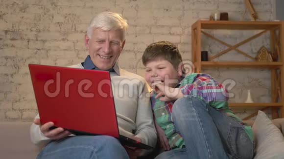 祖父和孙子坐在沙发上在笔记本电脑上看着一部有趣的电影笑着家庭舒适视频的预览图