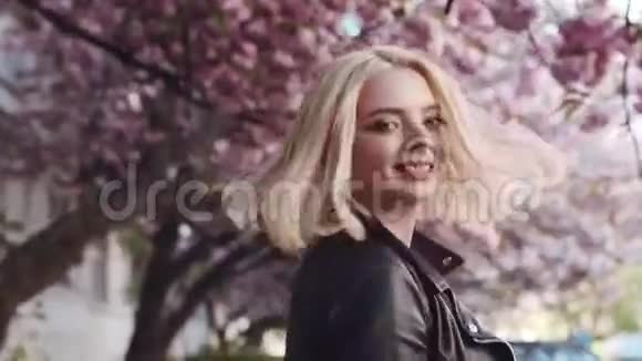 在樱花盛开的城市中漫步的迷人的金发少女的后景转向镜头微笑视频的预览图