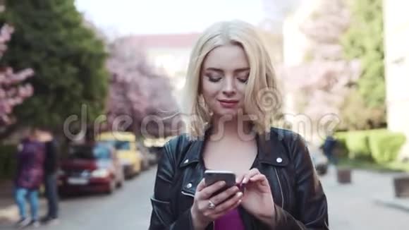 美丽的女孩穿着粉红色的上衣和皮夹克漫步在樱花盛开的城市梦幻般地上网视频的预览图
