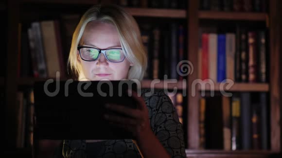一个戴眼镜的女人在书架的背景上用平板电脑工作上面有书概念图书馆工作或学习直至视频的预览图