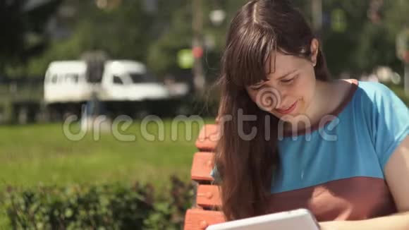 年轻的黑发女人在外面坐在长凳上用数码平板电脑视频的预览图