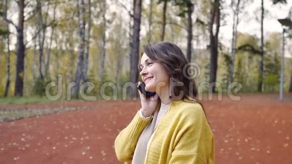 秋天公园里穿着一件带电话的黄色毛衣的年轻女人公园里的女人用手机打电话女孩视频的预览图