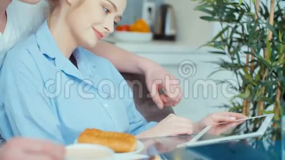 年轻夫妇在厨房餐桌上吃早餐时使用数字平板电脑视频的预览图