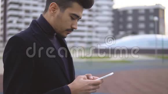 年轻的城市商人穿着黑色西装在街上行走在智能手机上使用应用程序发短信视频的预览图