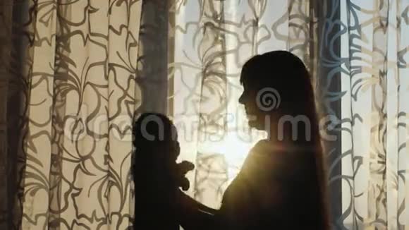 日落时分一位年轻的母亲在窗边和婴儿玩耍的剪影视频的预览图