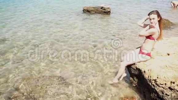 穿红色泳衣的女孩坐在岩石上双脚在水里视频的预览图