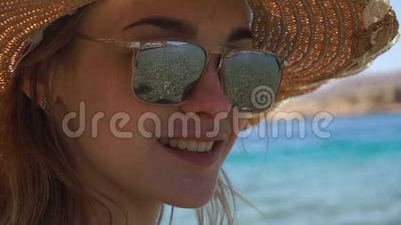 一个年轻的女孩戴着一顶帽子和一副眼镜你可以透过它看到大海看着相机微笑着视频的预览图